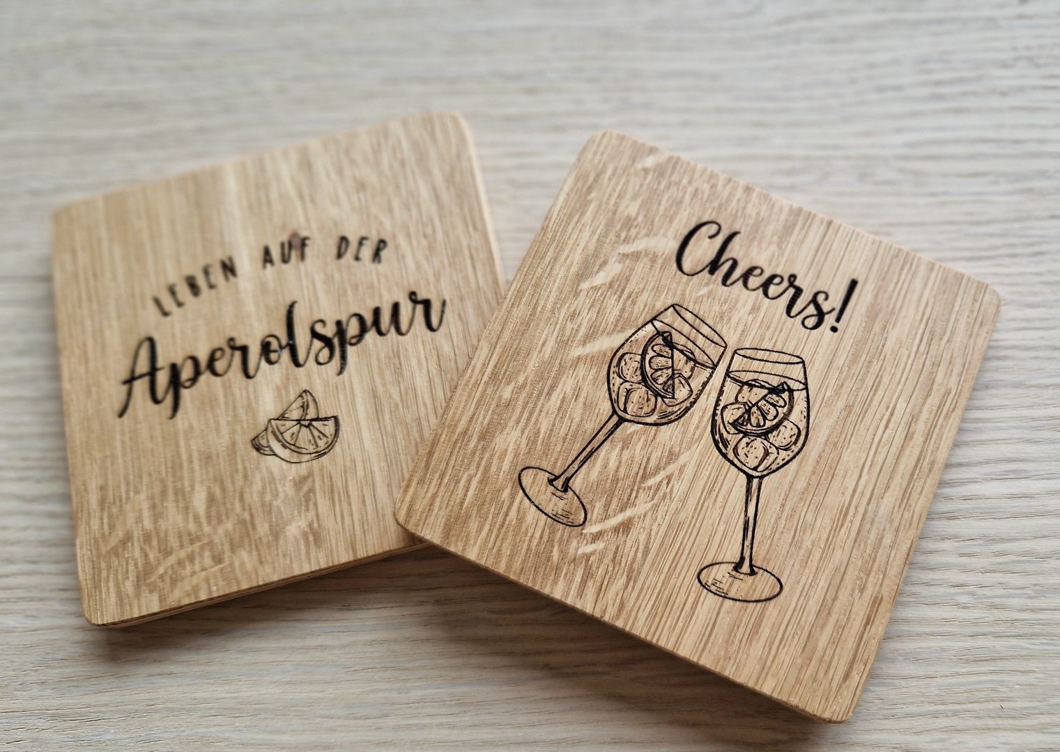 Zwei quadratische Holzuntersetzer mit Gravuren, einer mit der Aufschrift Leben auf der Aperolspur und einer mit Cheers! sowie zwei gravierten Weingläsern.